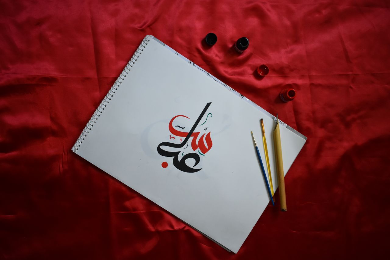 A top-down view of Arabic calligraphy on paper, styled text, and pens on red fabric.