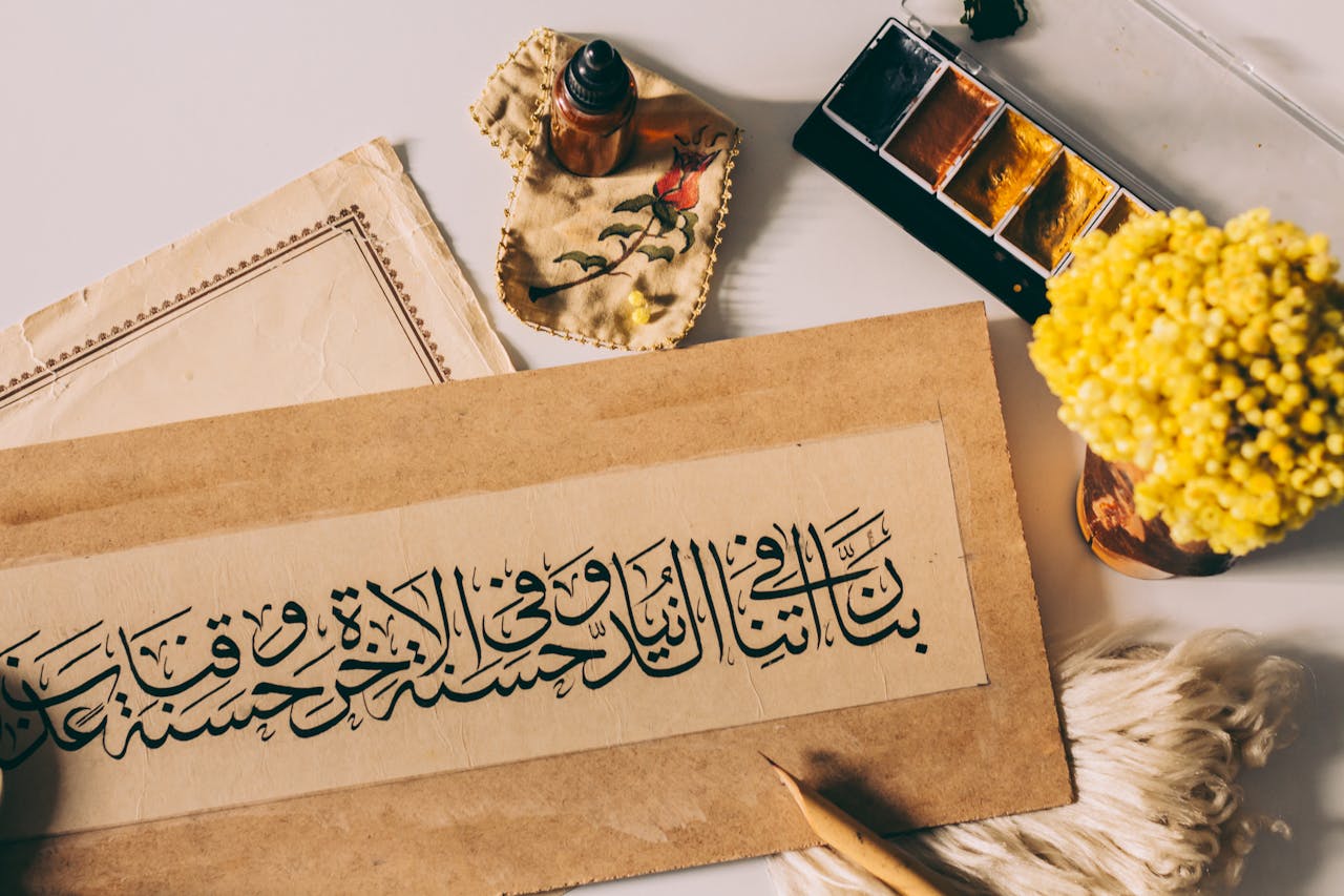 Artistic arrangement featuring Arabic calligraphy, paint, and flowers on a warm-toned workspace.